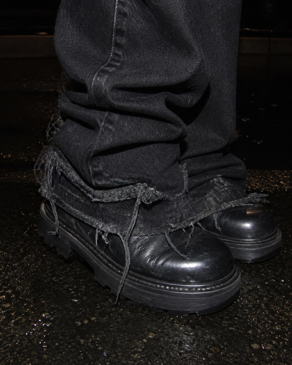 Close crop of a frayed black denim hem stacked over black boots on wet pavement