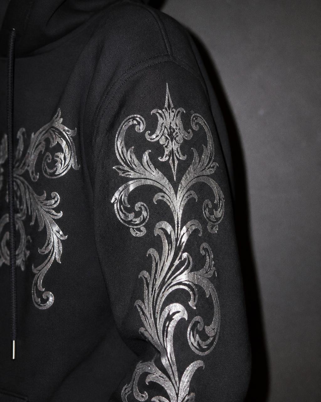 Shoulder and sleeve detail of a black hoodie with silver baroque print